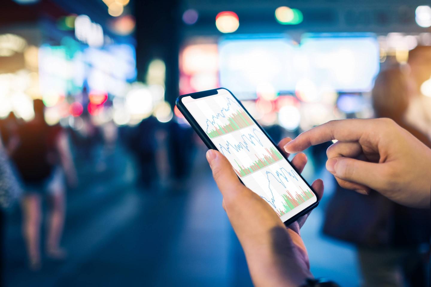 Close up of human hands checking financial stock charts on smartphone in busy city street, against neon commercial sign at night