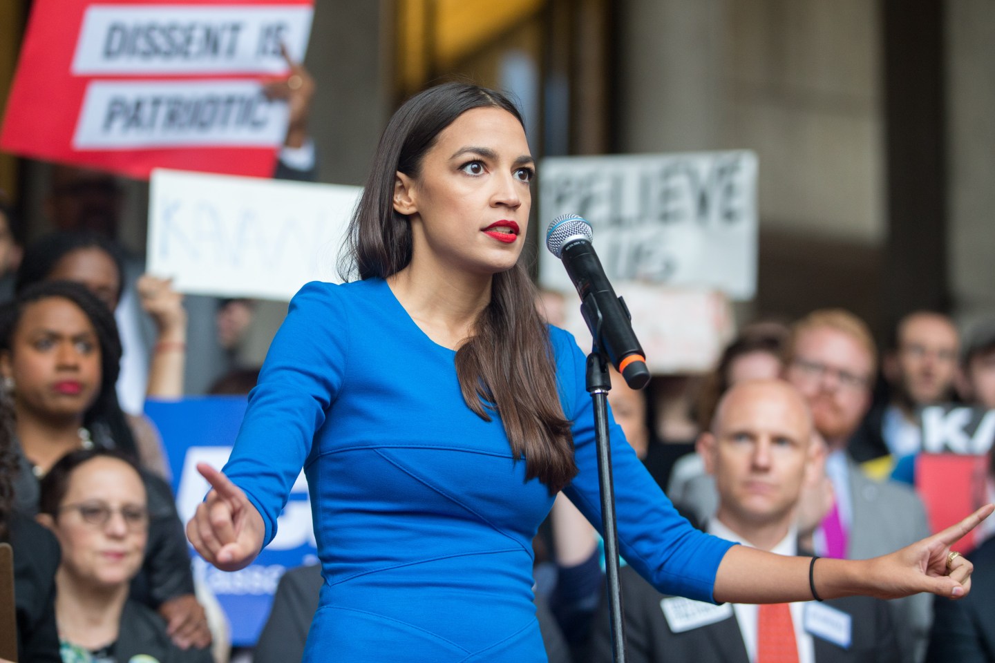 Alexandria Ocasio-Cortez stands in front of a microphone, speaking to a crowd in Boston.