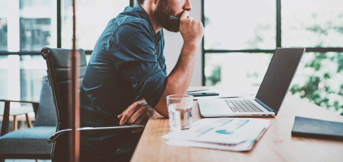 Young pensive coworker working at sunny work place loft while sitting at the wooden table.Man analyze document on laptop display.Blurred background.Horizontal wide.