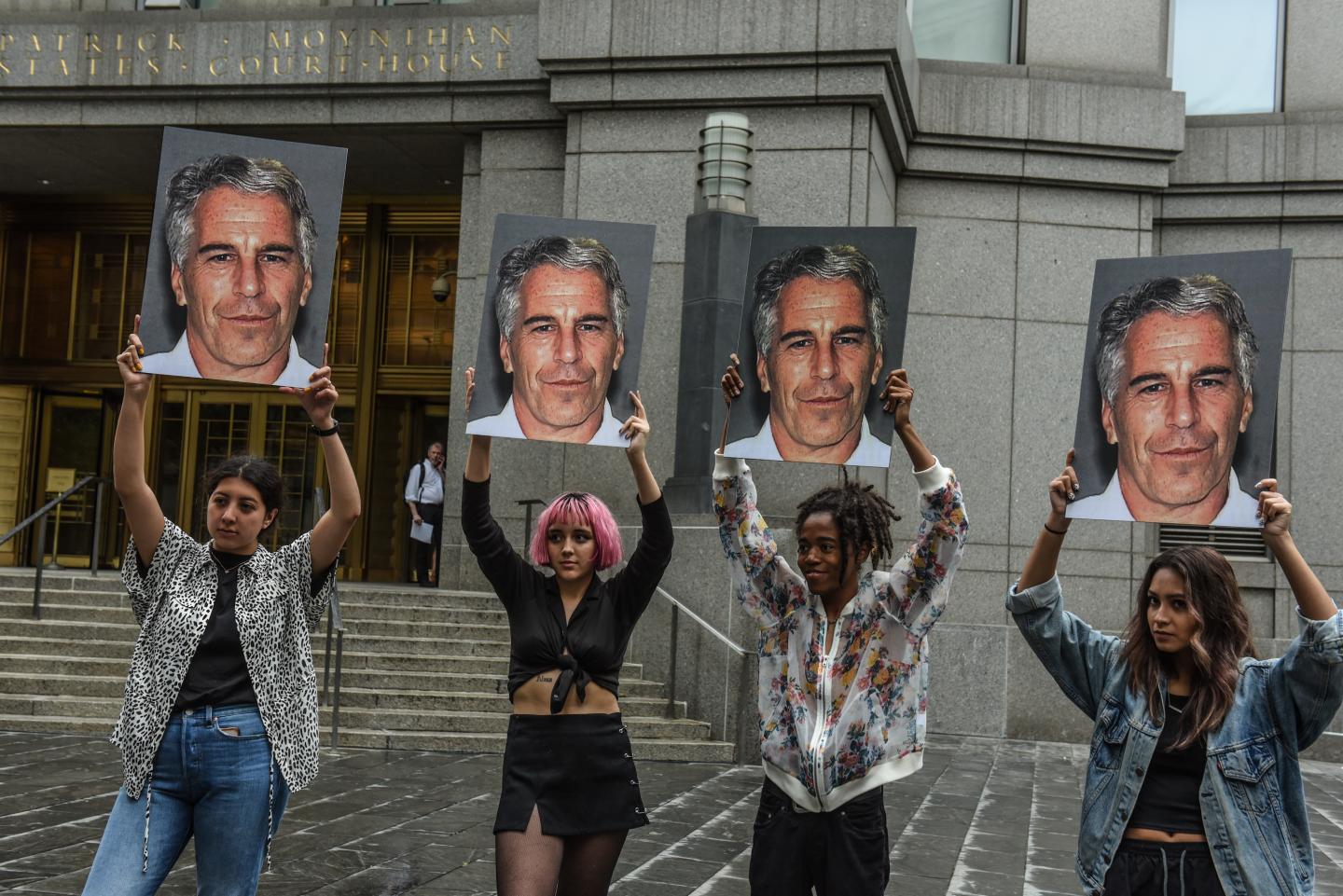 Four young women hold up signs with Jeffrey Epstein's mugshot on them.