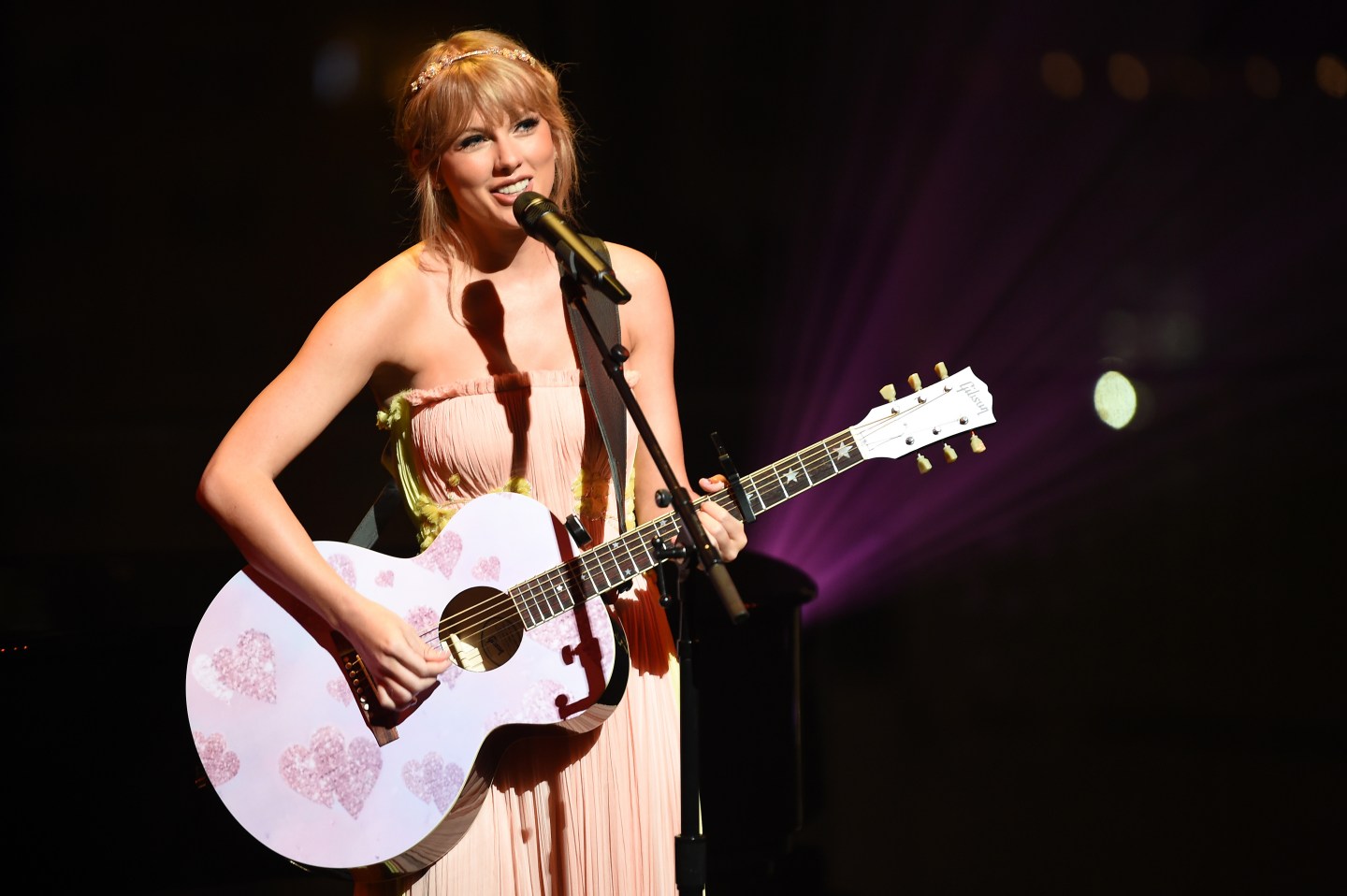Taylor Swift playing guitar and singing into a microphone against a black background