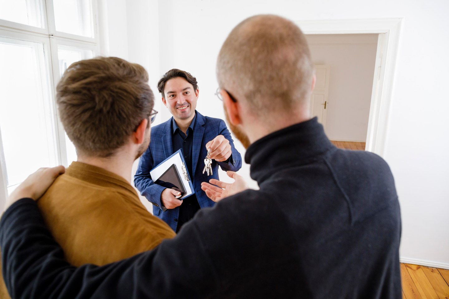 One man, facing the camera, hands a pair of keys to a couple. The couple consists of one man in a black shirt with his arm around another man in a brown jacket.