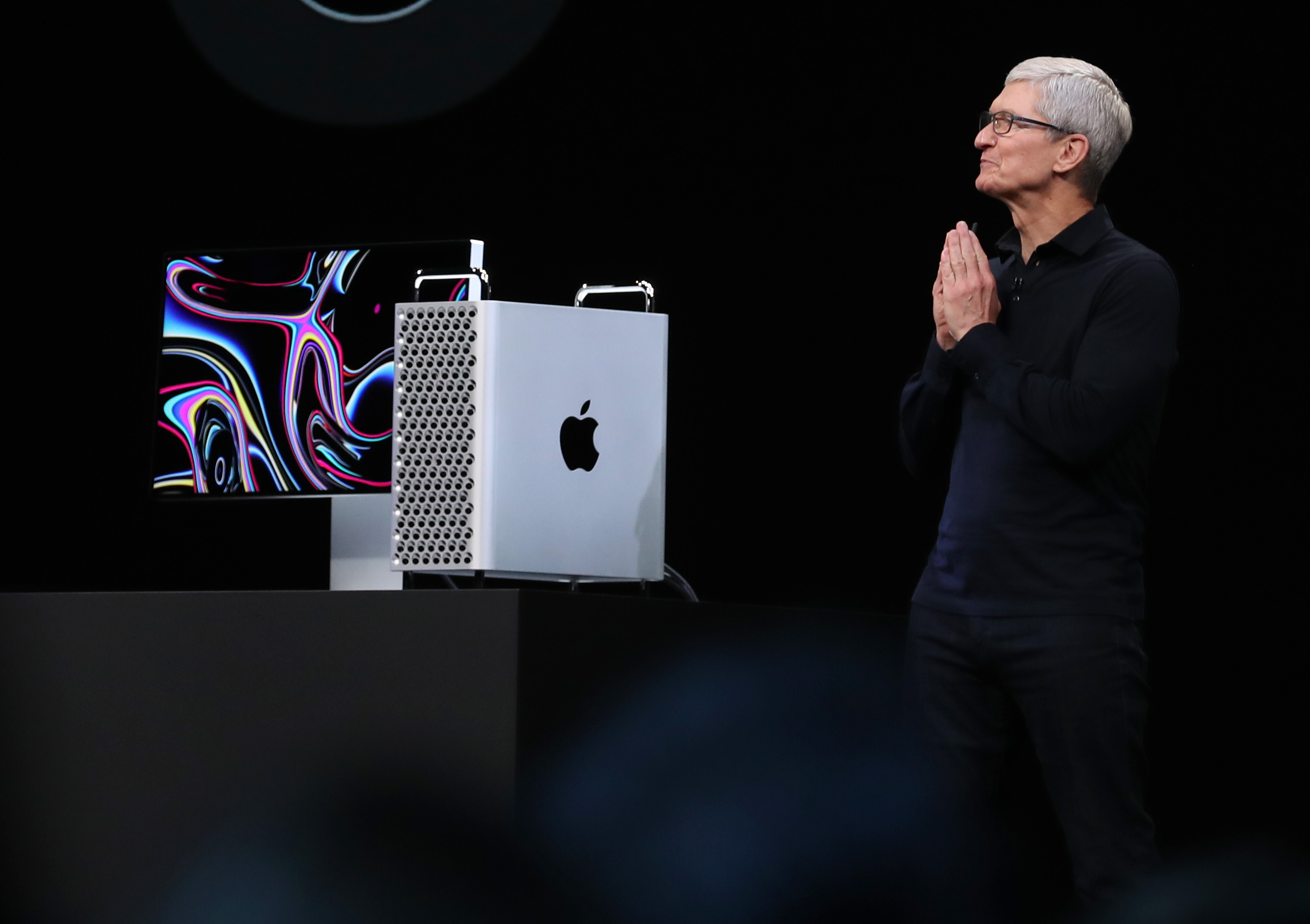 Tim Cook stands with hands pressed together next to the new Mac Pro, against a black background.
