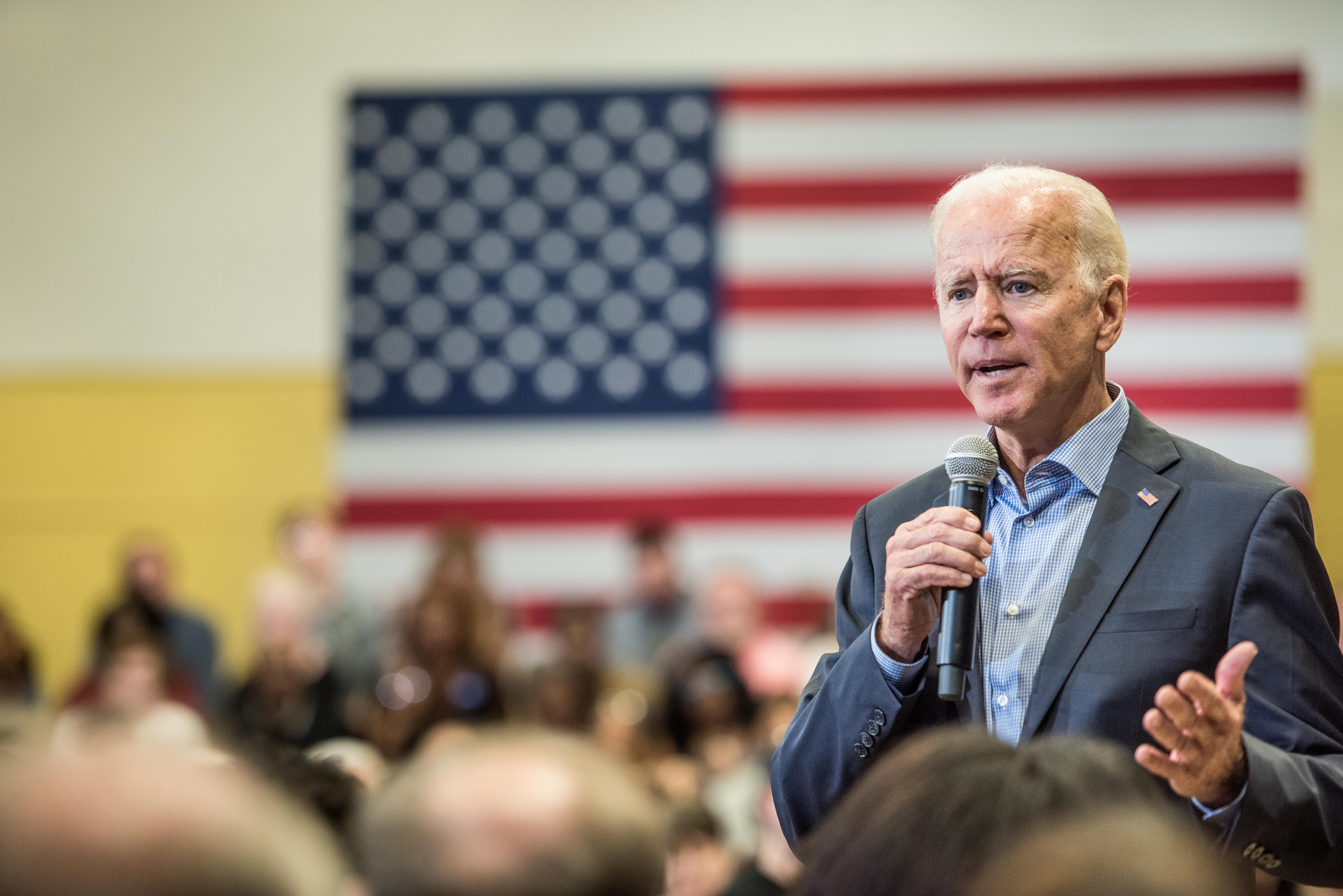 Joe Biden speaks into a microphone in front of a crowd with an American flag in the background.