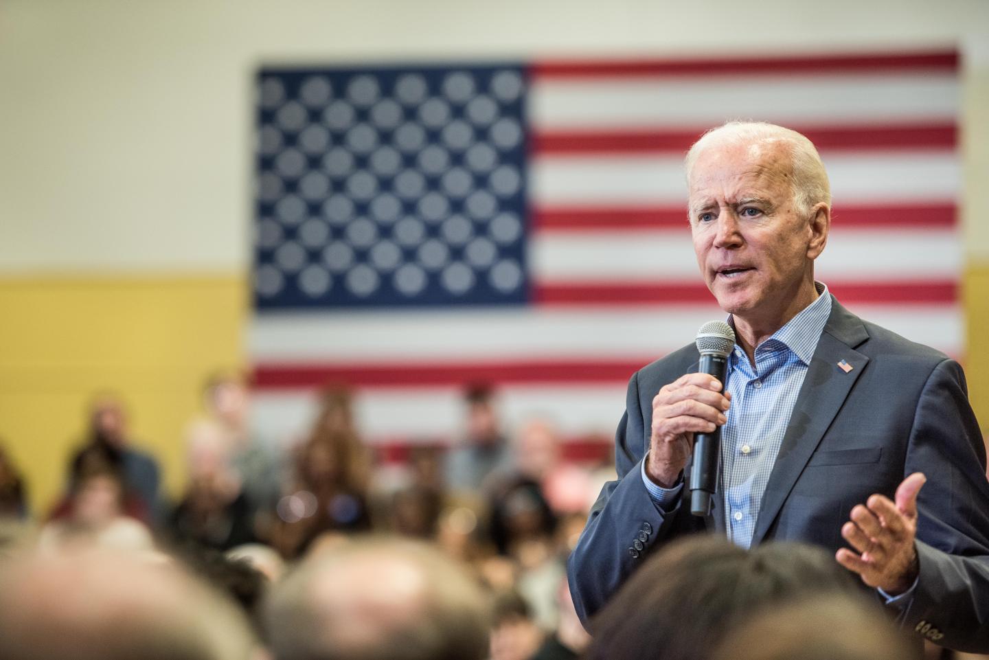 Joe Biden speaks into a microphone in front of a crowd with an American flag in the background.
