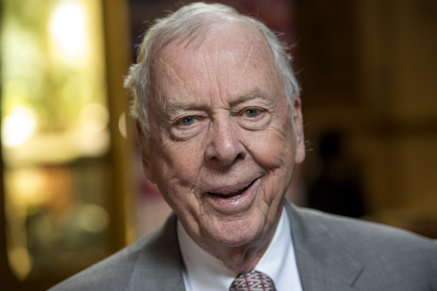 T. Boone Pickens smiles at the camera in a grey suit, white shirt, and patterned tie against a blurred, neutral background.