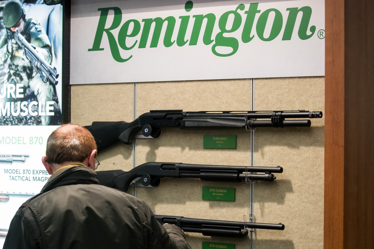 A Remington logo in green text on a white background hangs over a display of three long, black rifles. A man looks at the display.