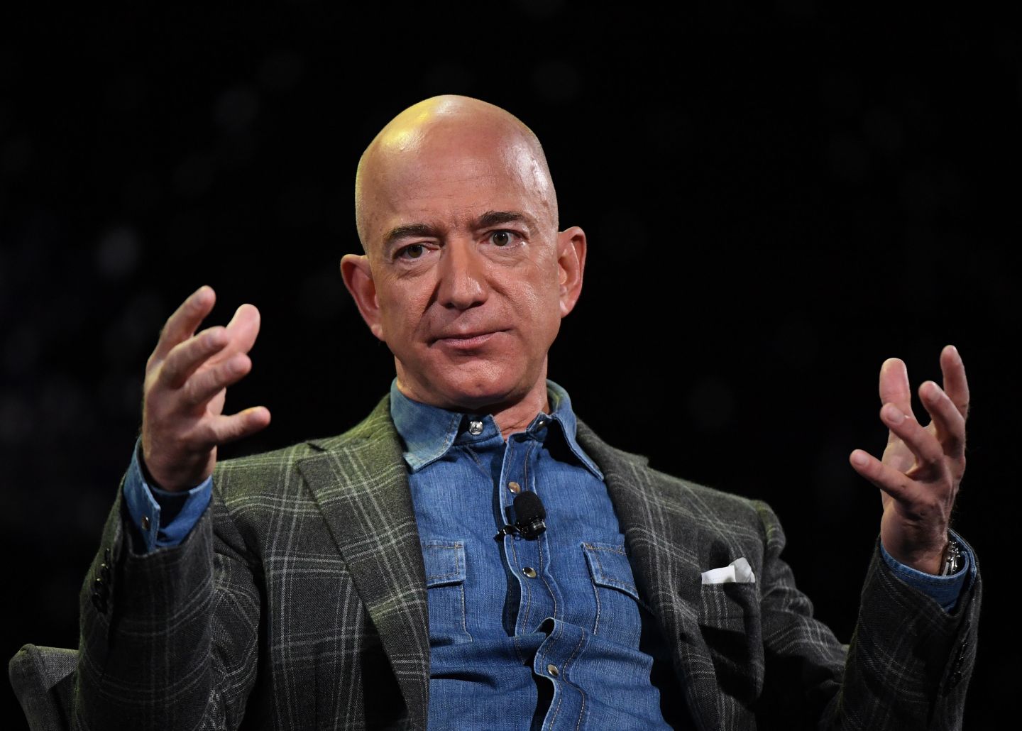 Jeff Bezos has both hands raised in front of him, looking frustrated. He is wearing a denim shirt and a plaid blazer.