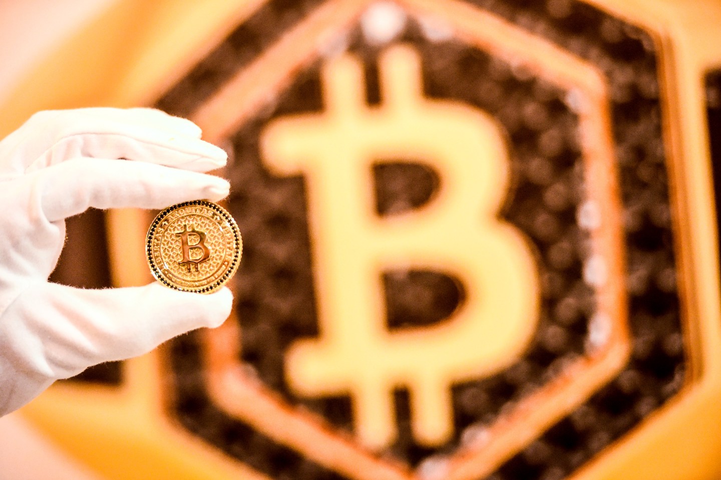 A white-gloved hand holds a Bitcoin coin in front of a hexagonal Bitcoin logo, blurred out in the background.