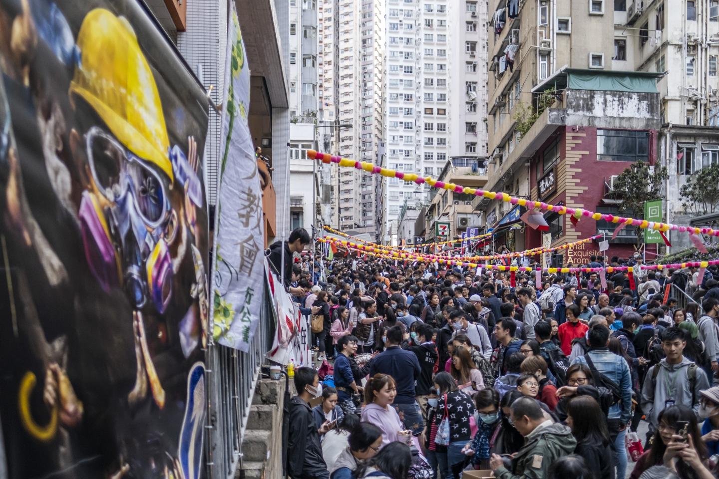 People walk past a protest banner during a Lunar New Year fair organized by pro-democracy district councilors on Centre Street in the Sai Ying Pun district of Hong Kong, China, on Saturday, Jan. 18, 2020. The Lunar New Year, when spending traditionally peaks in Hong Kong amid a flood of vacationing Chinese mainlanders, will likely not provide its usual boost after months of fiery demonstrations that have driven tourists away. Photographer: Justin Chin/Bloomberg via Getty Images