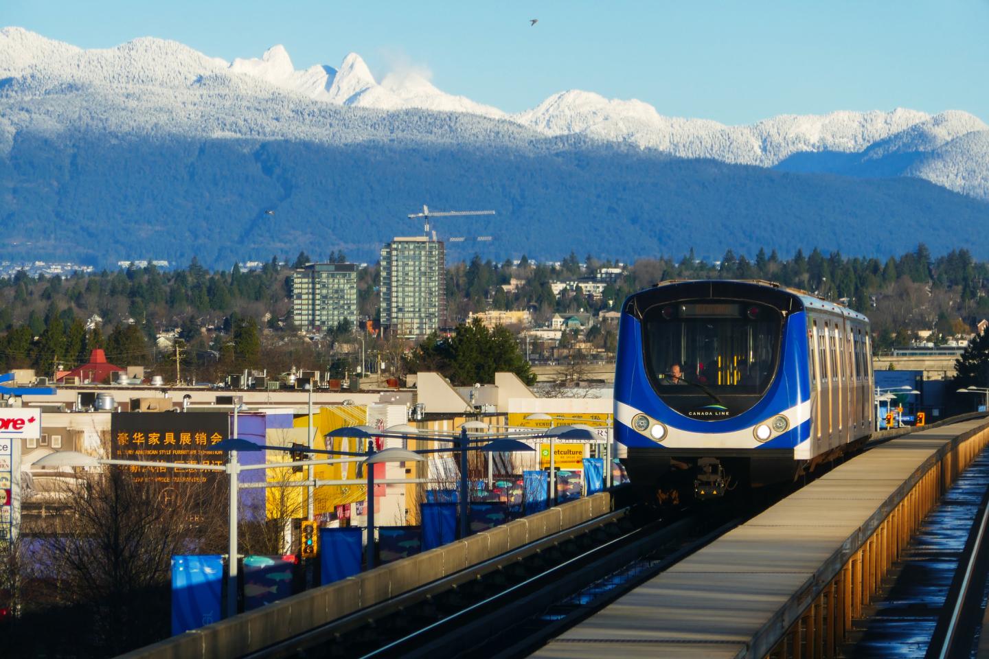 In winter, the Canada Line rapid transit on the elevated railway with city streets and buildings views and snow capped mountains as backdrop, between Richmond and Vancouver cities, in British Columbia, Canada.
