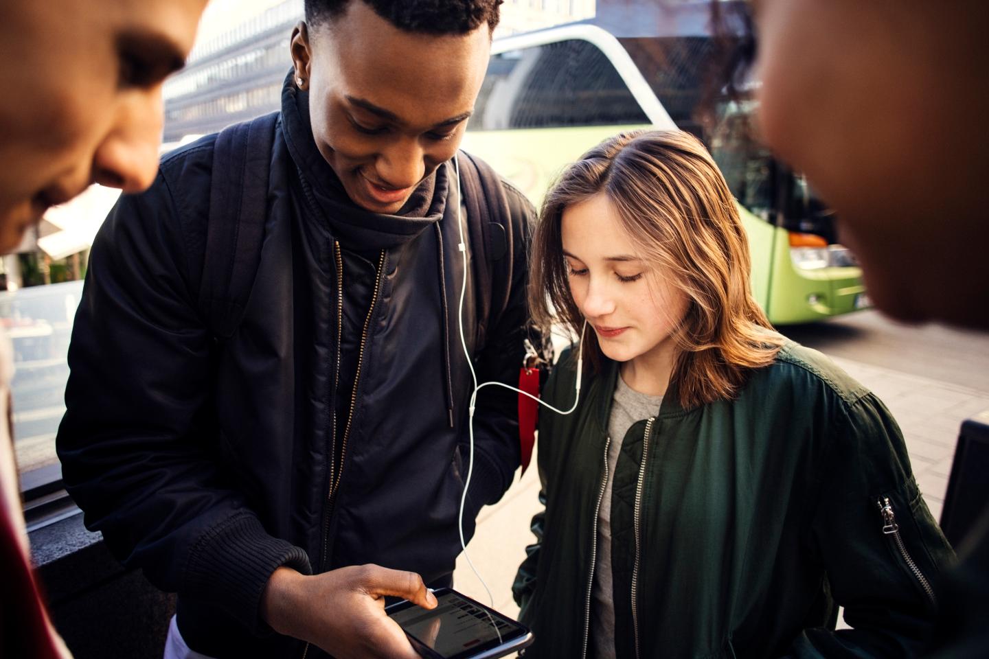 Teenage girl and boy listening music from smart phone while showing to friends in city