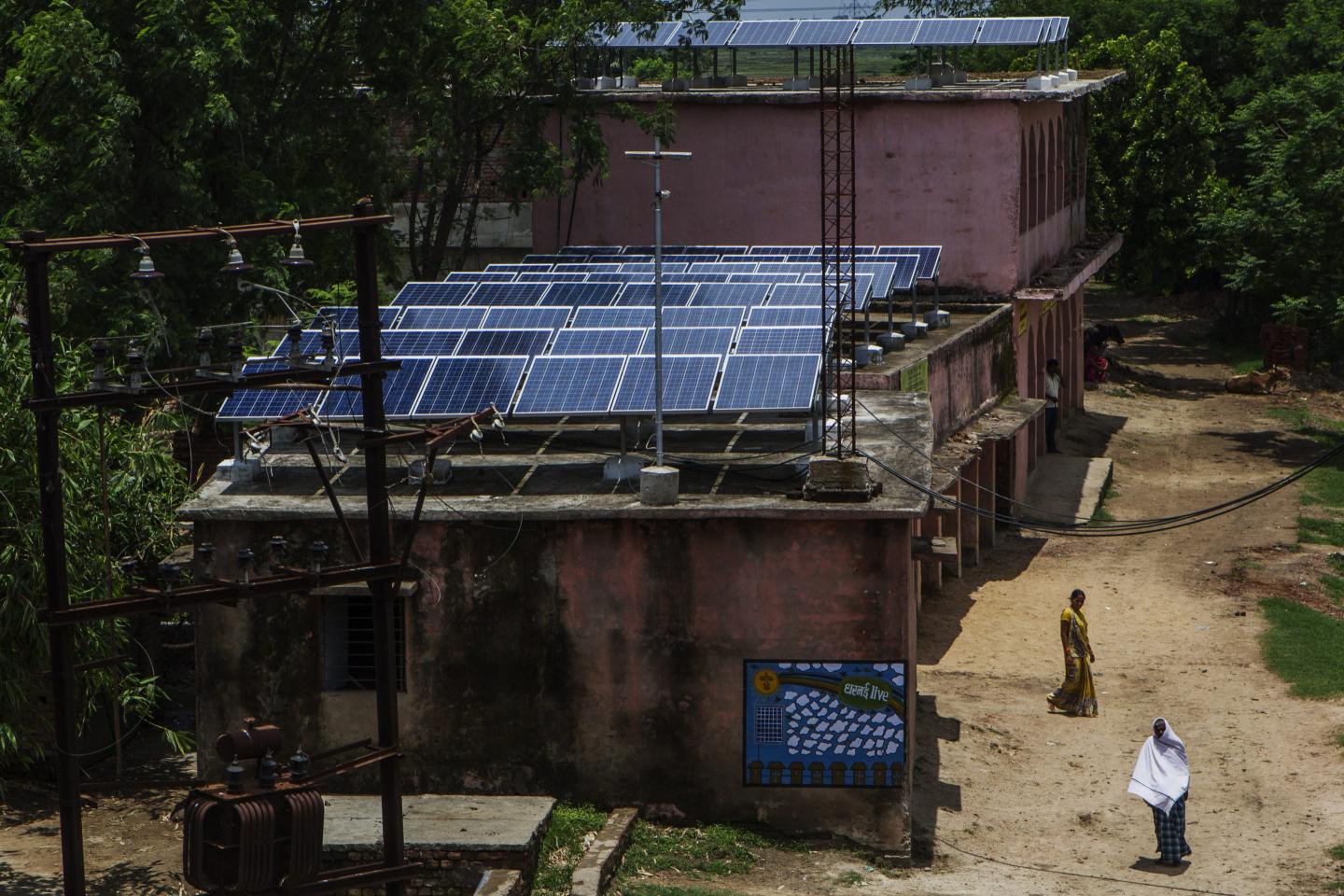 Villagers walk past solar panels mounted on the roof of a building, part of a solar power microgrid, in the village of Dharnai in Jehanabad, Bihar, India, on Thursday, July 9, 2015.