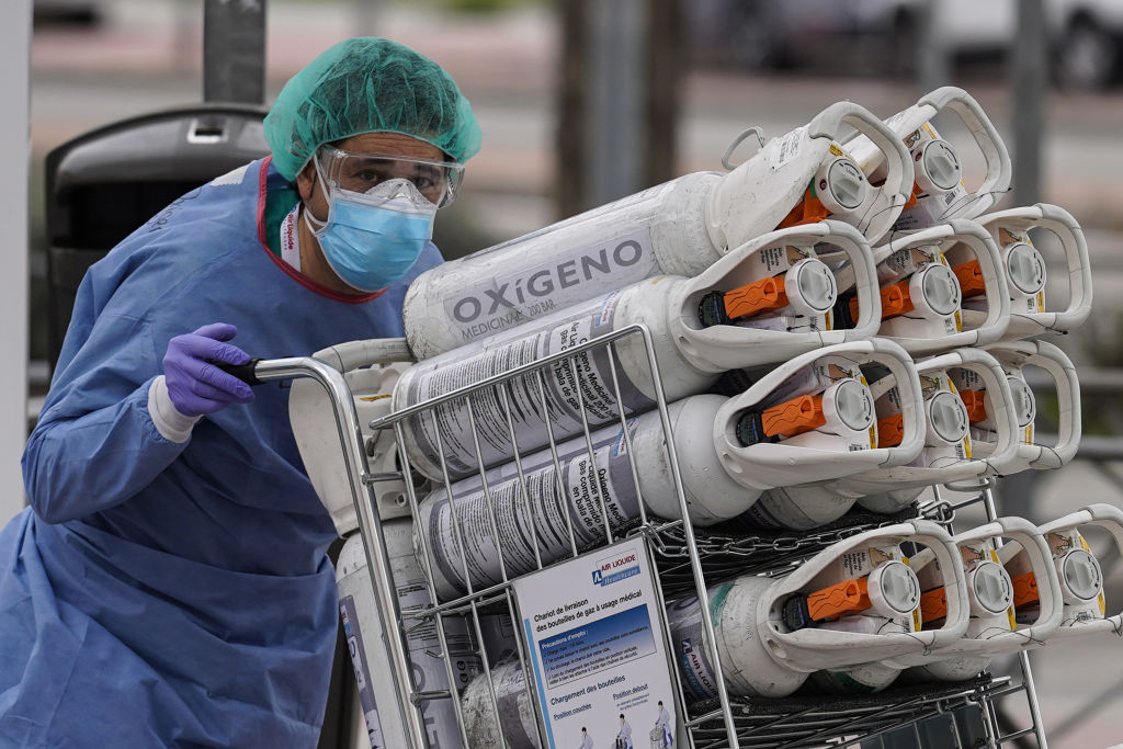 Medical worker in scrubs and mask pushing a cart full of oxygen tanks outside a hospital in Spain.