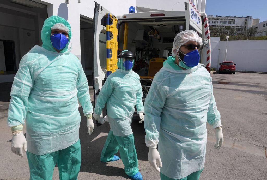 Three doctors in protective equipment walking outside with an ambulance in the background.