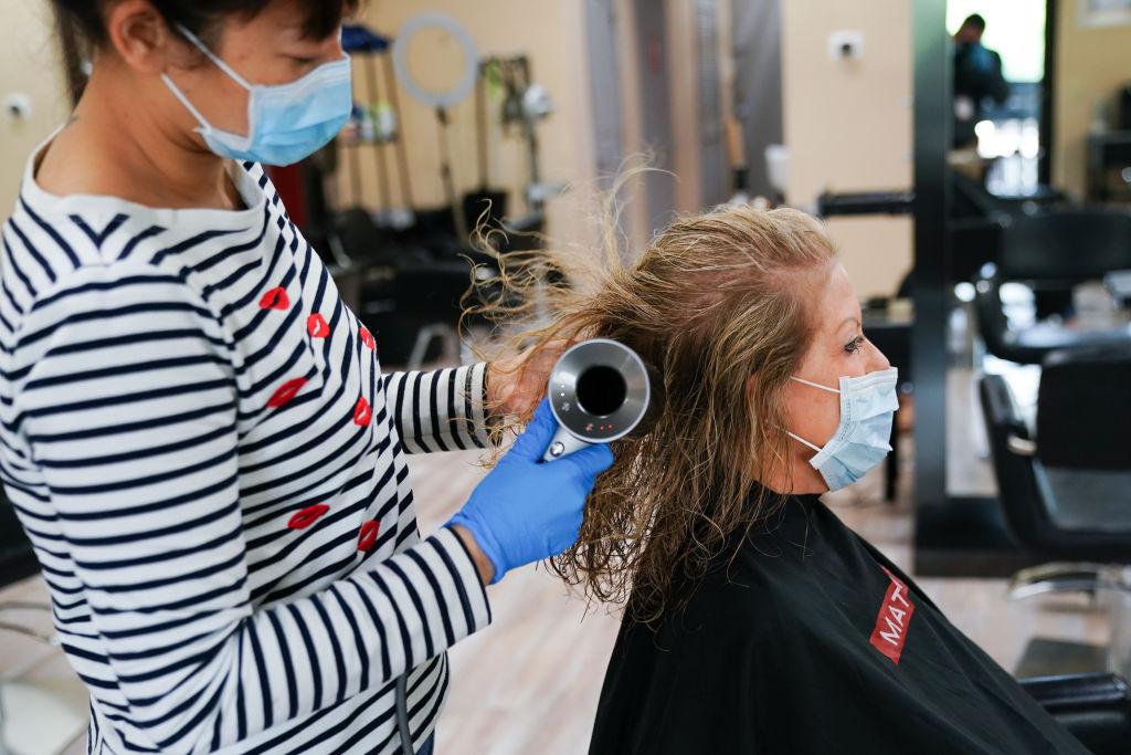 Hairdresser in face mask blow drying the hair of a woman who also wears a face mask.