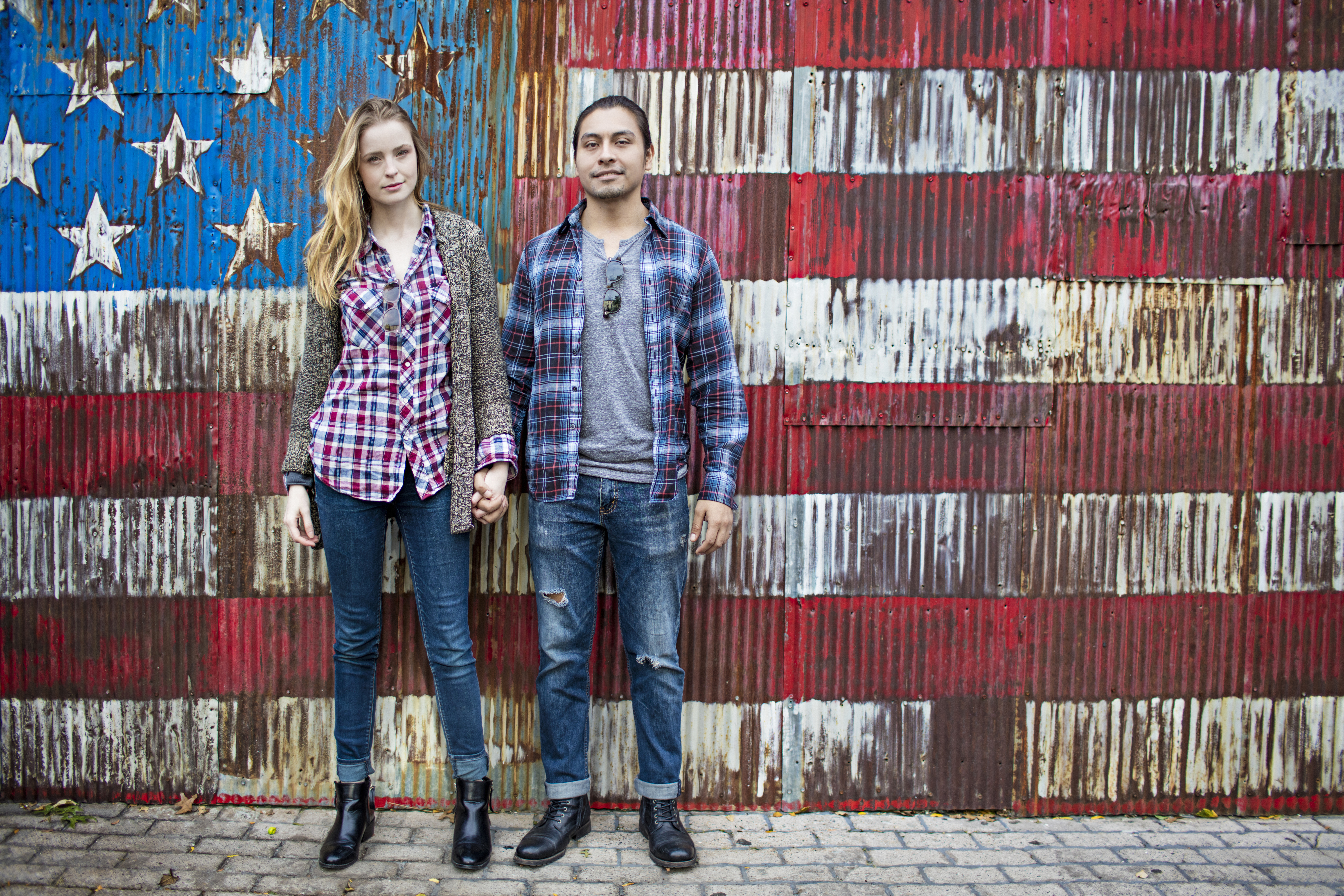 Full length portrait of confident couple standing against corrugated wall. Young man and woman are against American flag painted on rusty metal. They are in casuals.