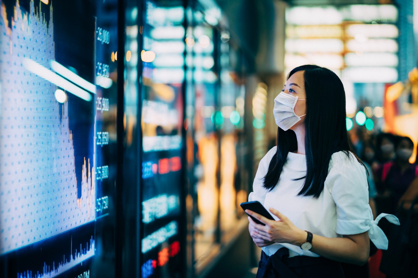 Woman wearing a face mask looking at a big screen while holding a mobile phone.