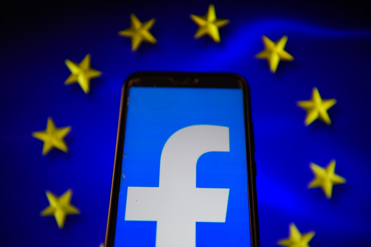 A Facebook logo is seen displayed on a smartphone with an European Union flag background.