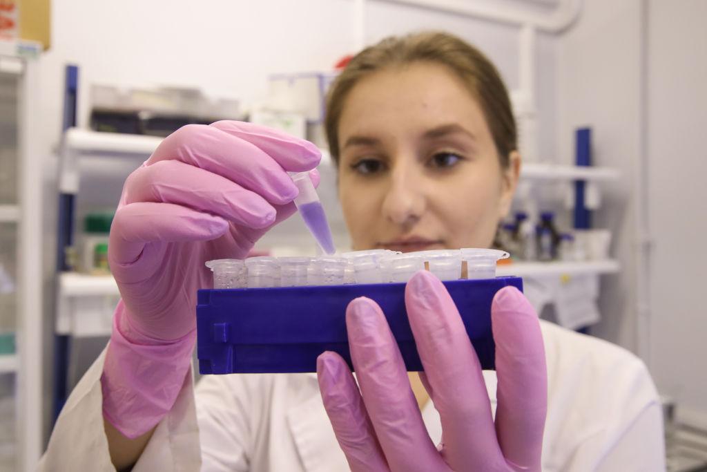 Woman in a white lab coat and pink rubber gloves holding a vial of purple liquid.
