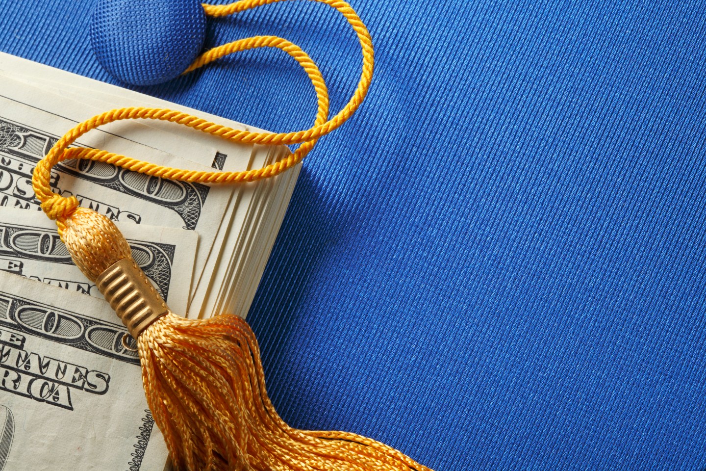 A stack of one hundred dollar bills on top of a blue graduation cap. A gold graduation tassel is draped over the stack of money.