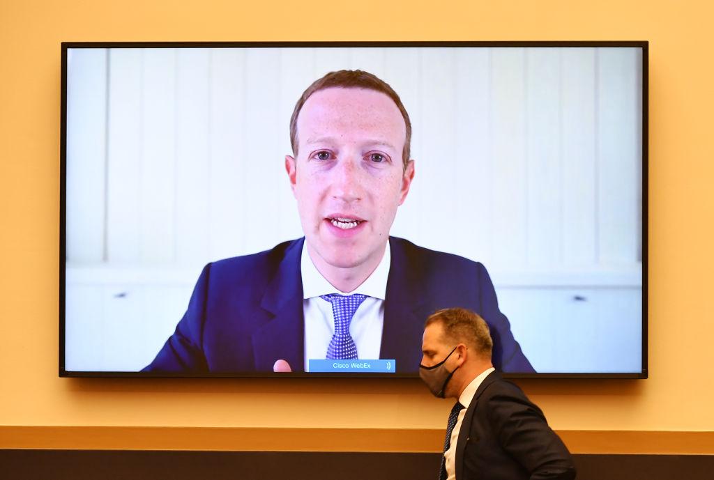 Photo of a big TV screen filled with an image of Facebook CEO Mark Zuckerberg testifying before Congress while a man in a suit and with a mask on walks in front of the screen.