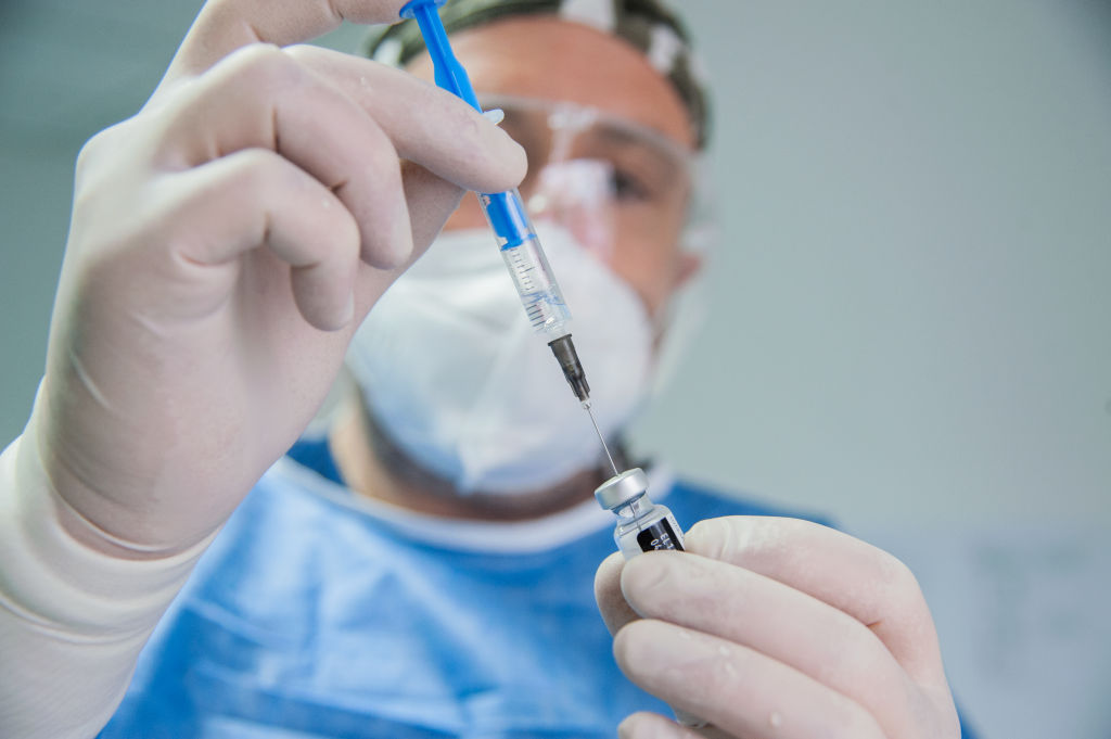 A medical professional in hat, mask, goggles and gloves draws a syringe of vaccine from a vial.