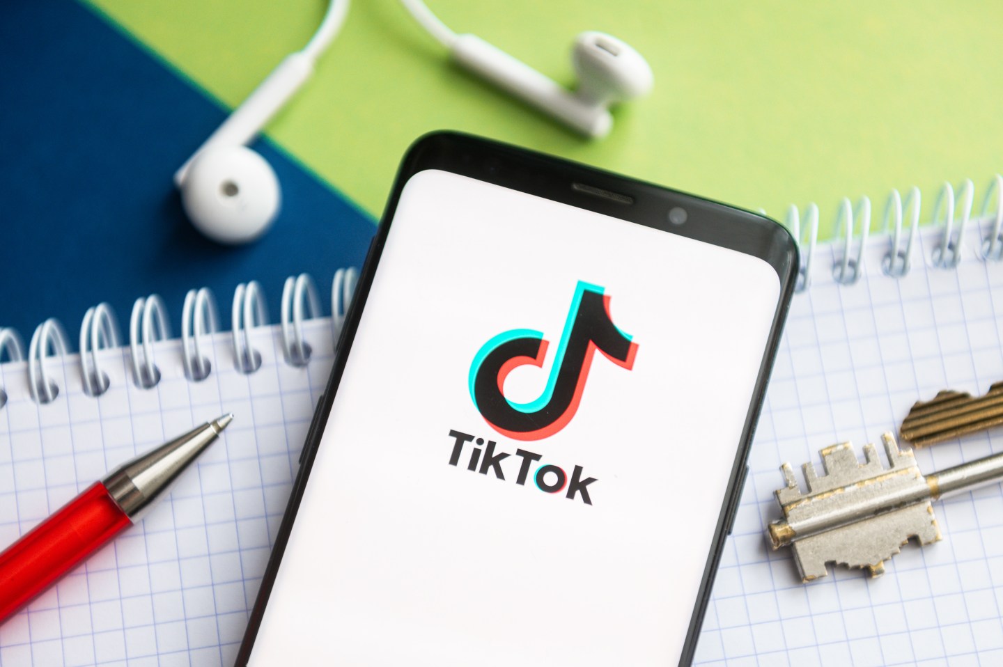 In this photo illustration, a Tik Tok logo seen displayed on a smartphone with a pen, key, book and headsets in the background.