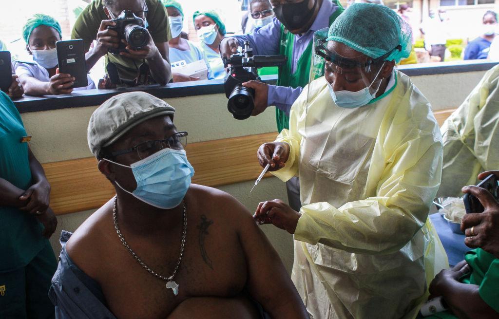 A man with his shirt off and wearing a mask receives a COVID-19 vaccination from a health worker wearing PPE.