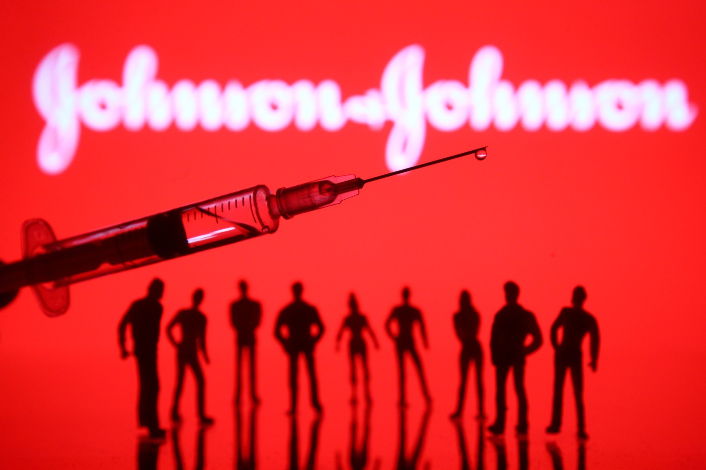 In this photo illustration a Johnson &amp; Johnson logo is seen in front of silhouettes people and a medical syringe.