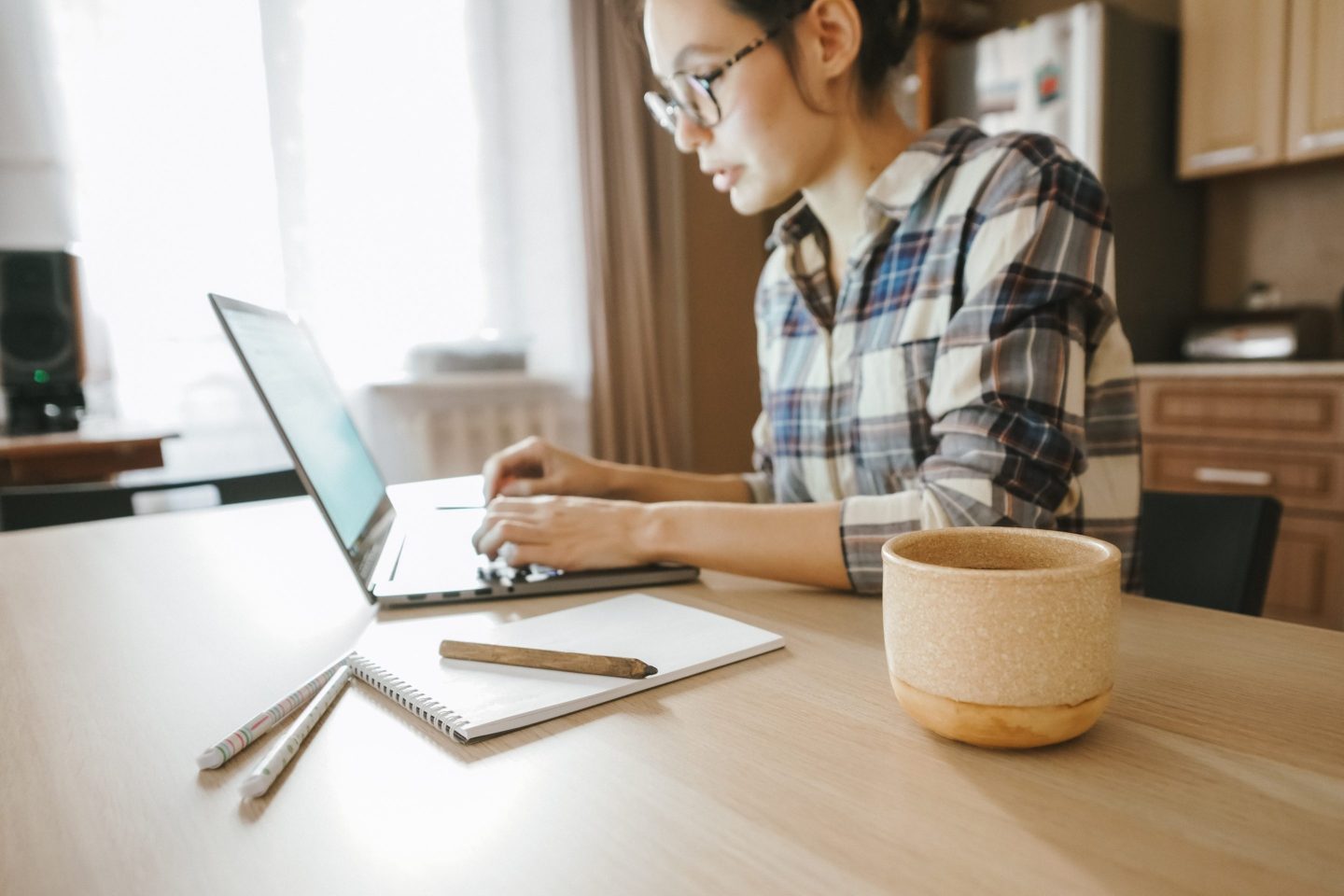 Woman sits at a table working on a laptop, with a coffee cup and notepad nearby.