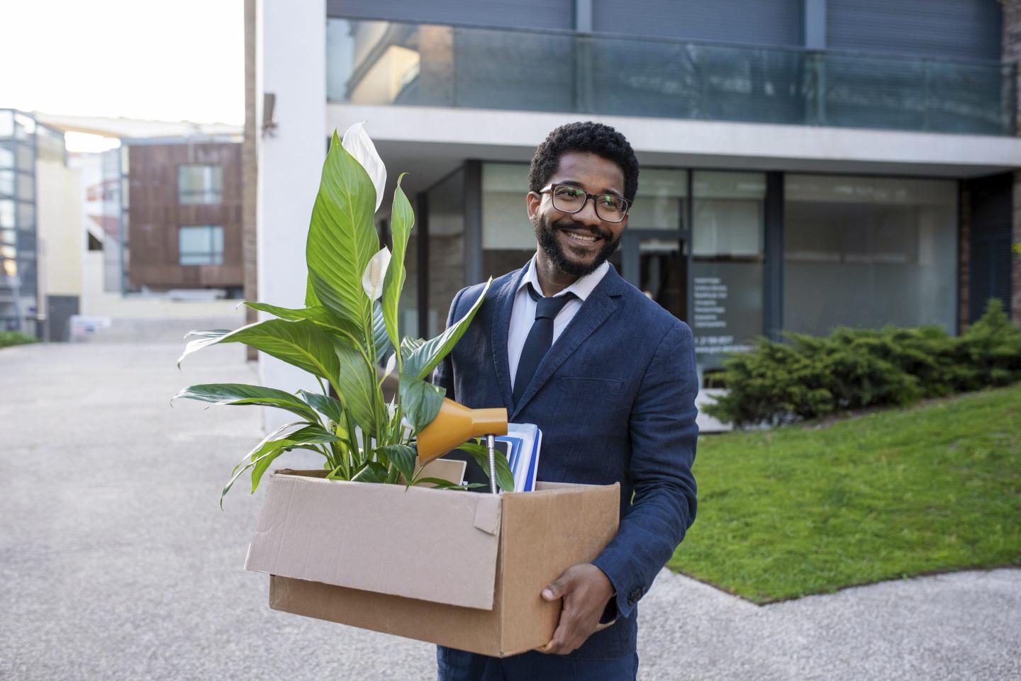 Businessman holds a box with a plant sticking out as he leaves an office.