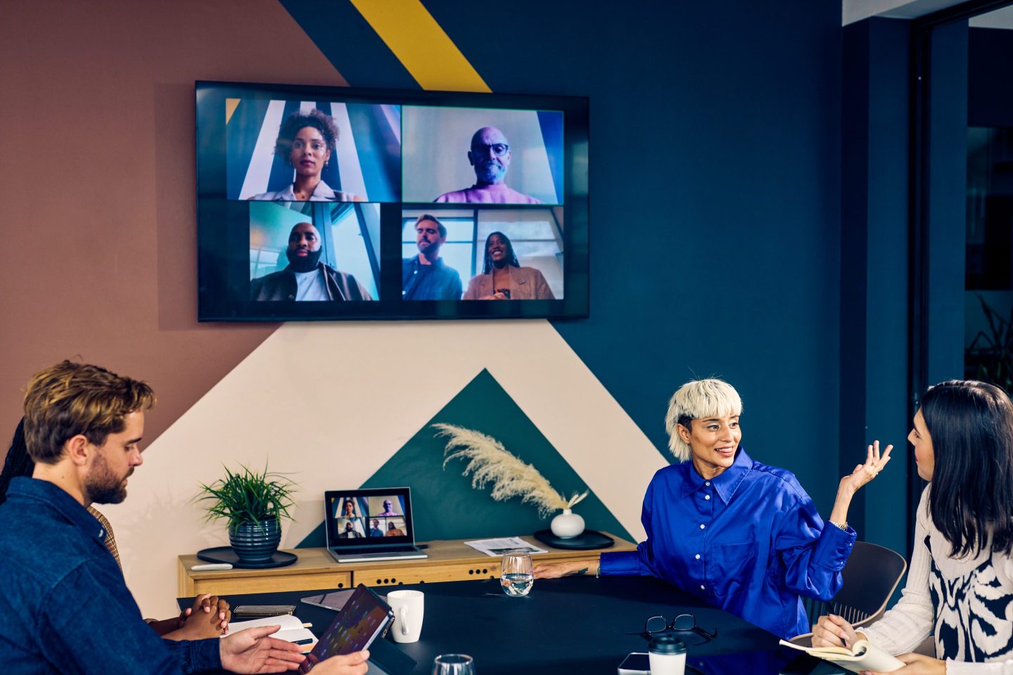 Students sit a table while a screen behind them shows virtual learners.