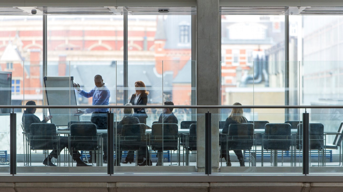Group of six business people in a boardroom meeting. Shot at a distance from outside through the glass.