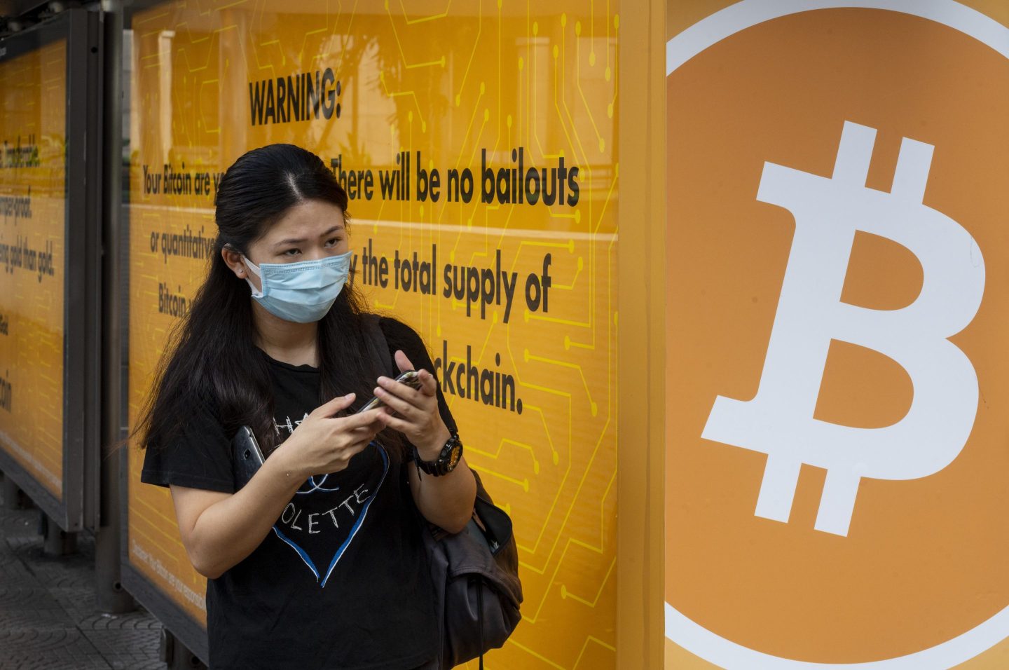 HONG KONG, CHINA &#8211; 2020/09/24: A woman wearing a mask stands next to a bus stop covered with Cryptocurrency electronic cash Bitcoin advertisement in Hong Kong. (Photo by Budrul Chukrut/SOPA Images/LightRocket via Getty Images)