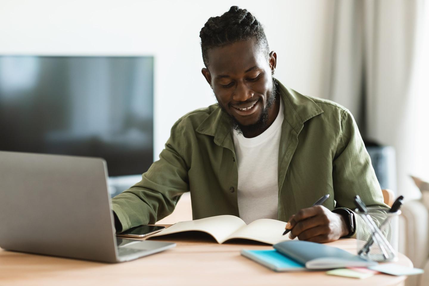 Man sits at a desk, writing in a notebook while working on his laptop.