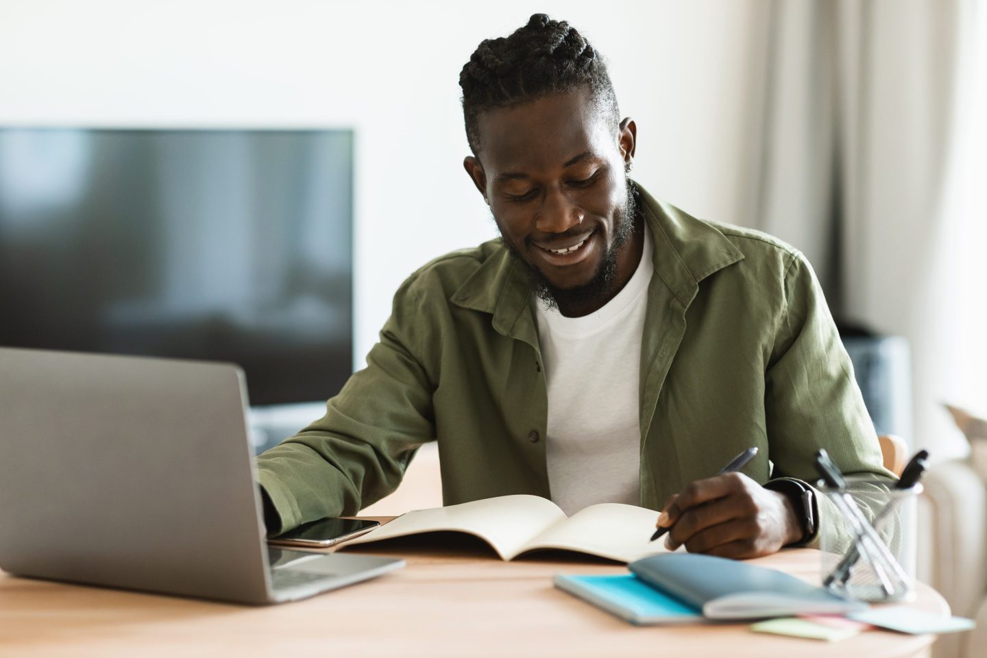 Man sits at a desk, writing in a notebook while working on his laptop.
