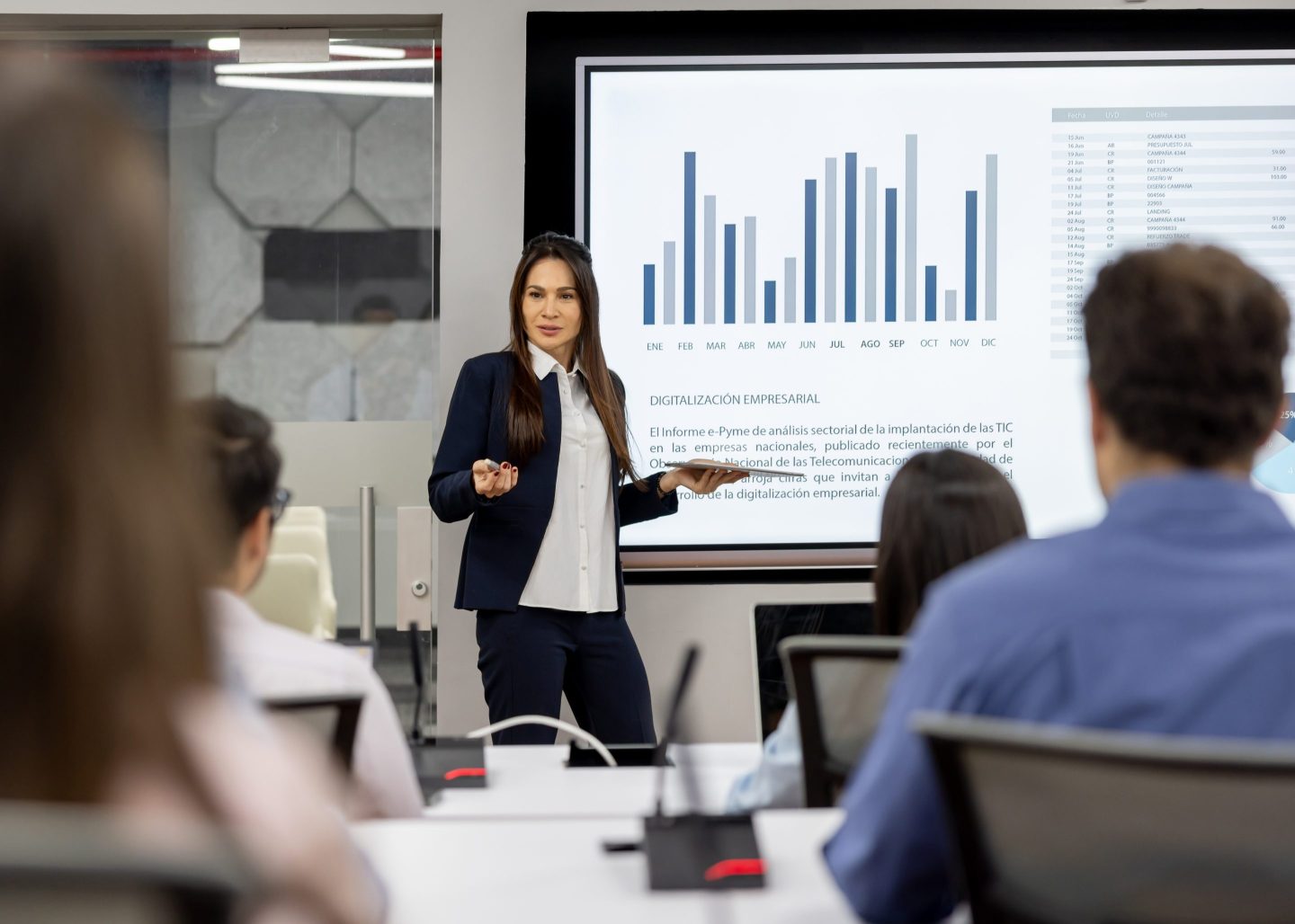 Woman stands in the front of a classroom teaching a group of adult students, with a chart behind her.