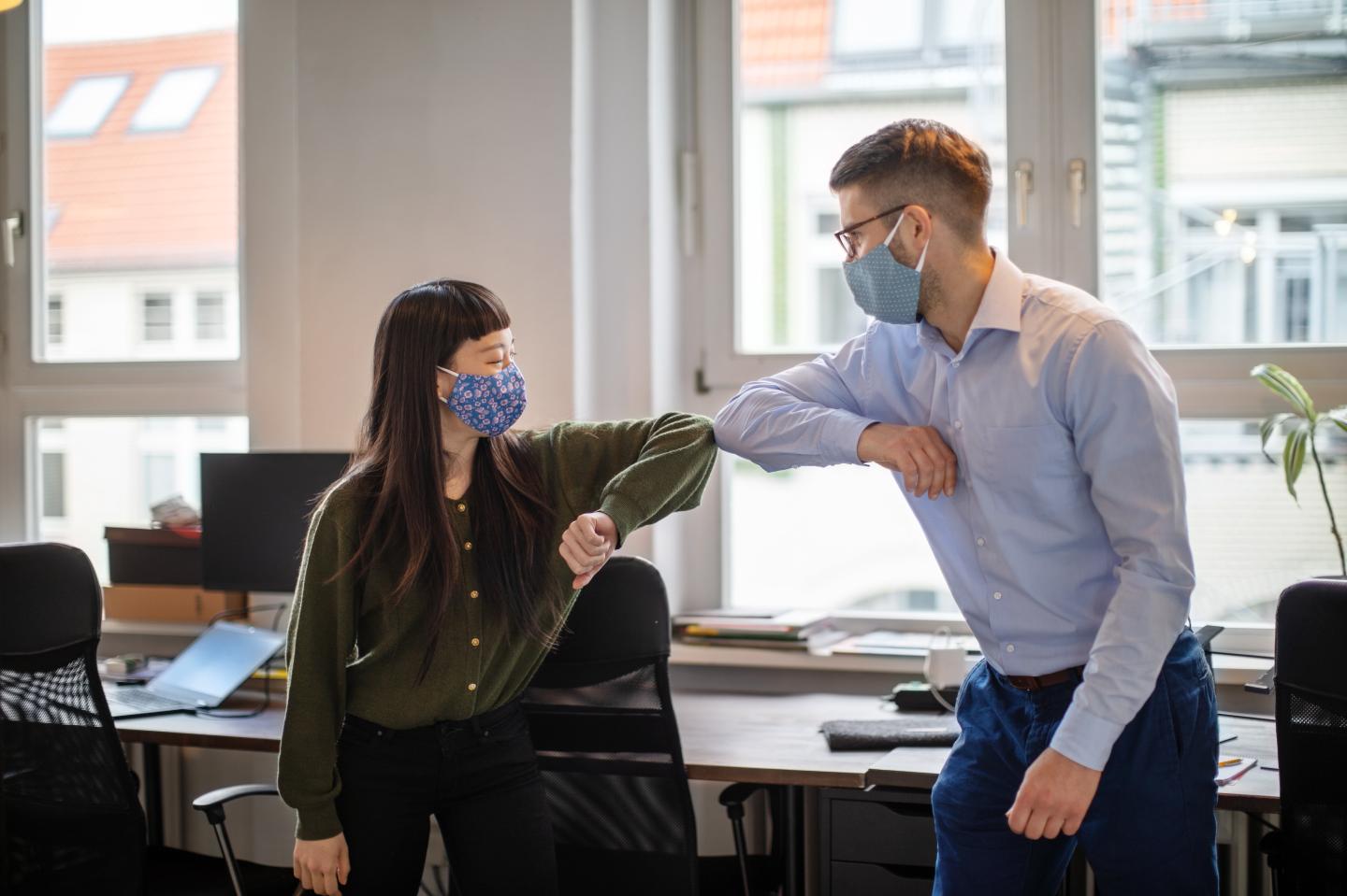 Business people in the office practicing alternative greeting for safety and protection during COVID-19. Two colleagues wearing safety face mask elbow bumping while greeting each other in the office.