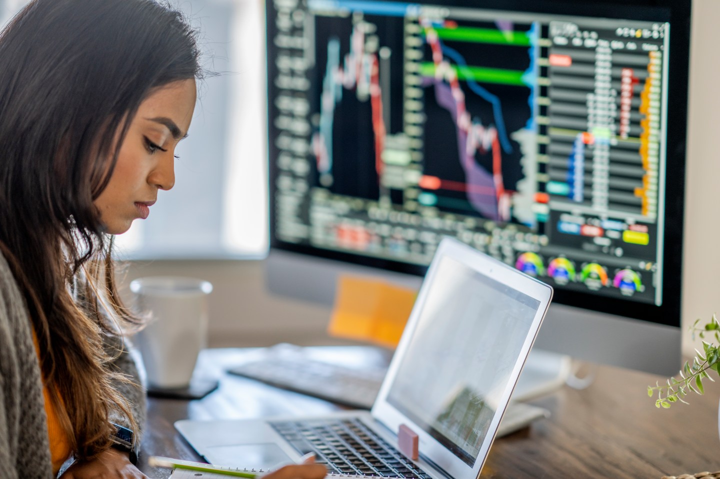 Middle Eastern woman tracking and trading stocks using laptop and desktop computer. Stock Exchange, investment, and financial trading concept.