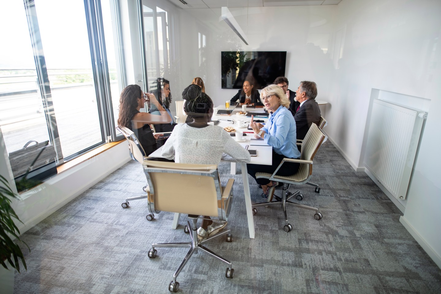 Wide angle view photographed through window of multi-ethnic group of corporate colleagues conversing at conference table in office board room.