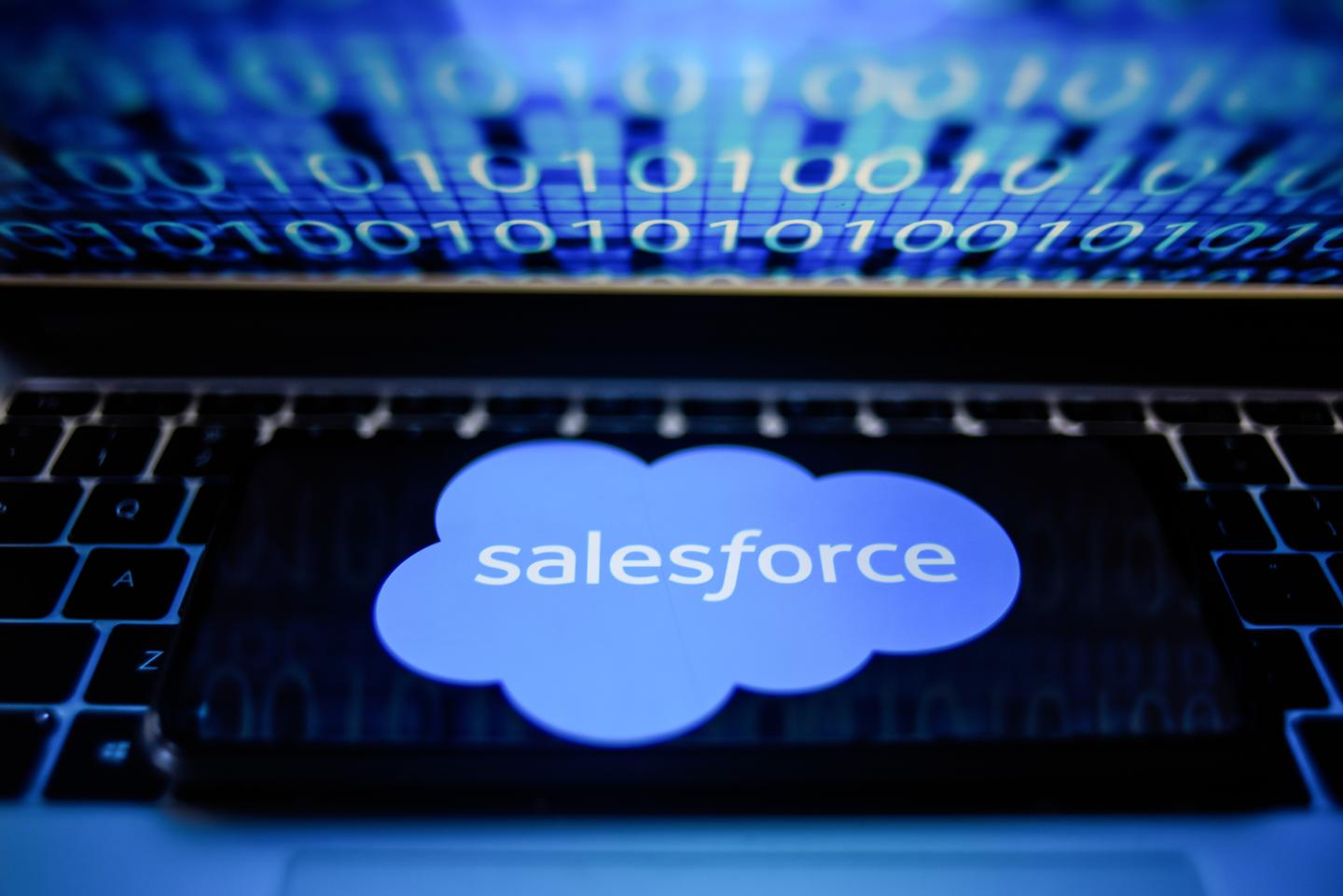In this photo illustration a SalesForce logo seen displayed on a smartphone with binary code on a laptop screen.