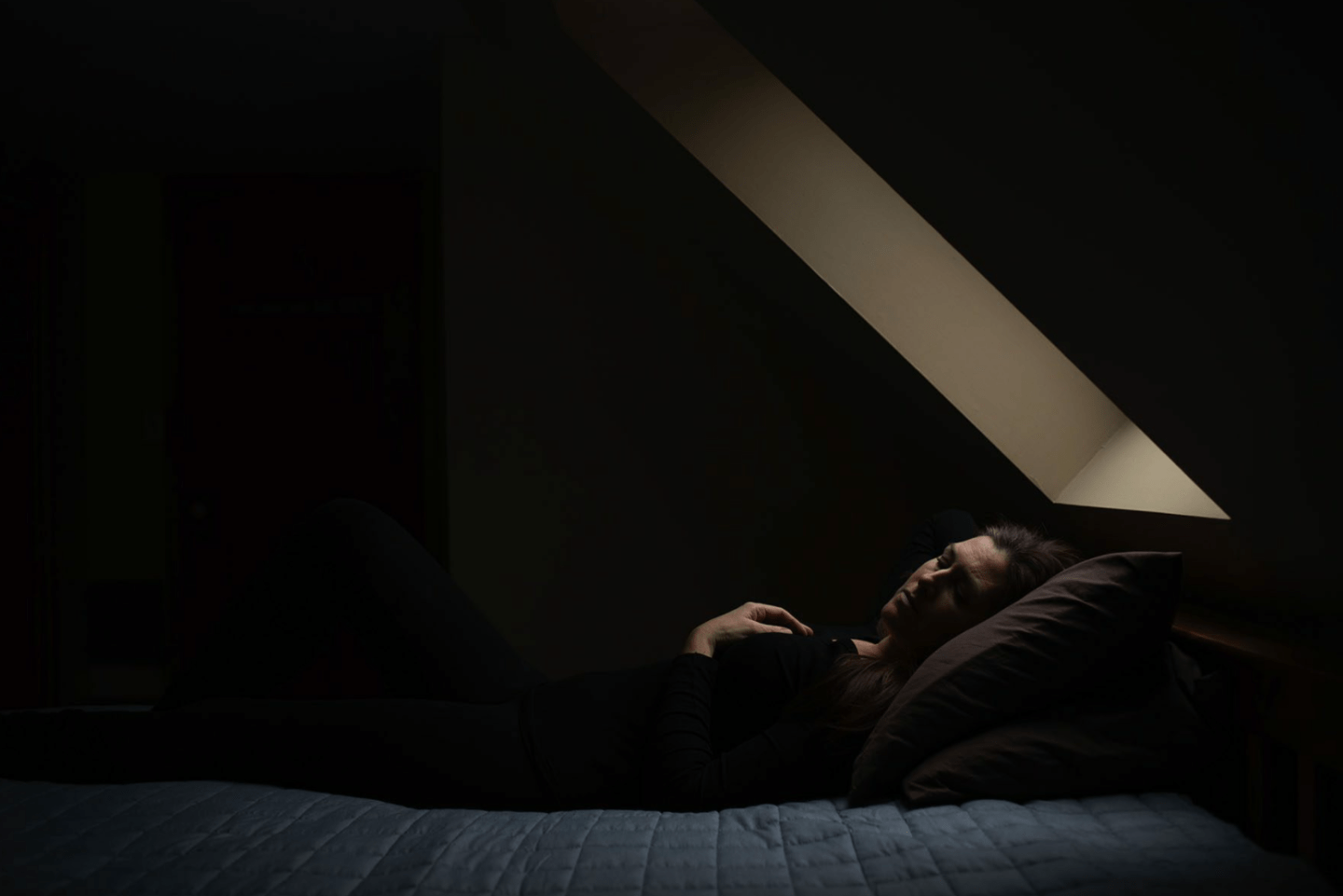Woman laying on bed with eyes closed in dark room under a sky light.