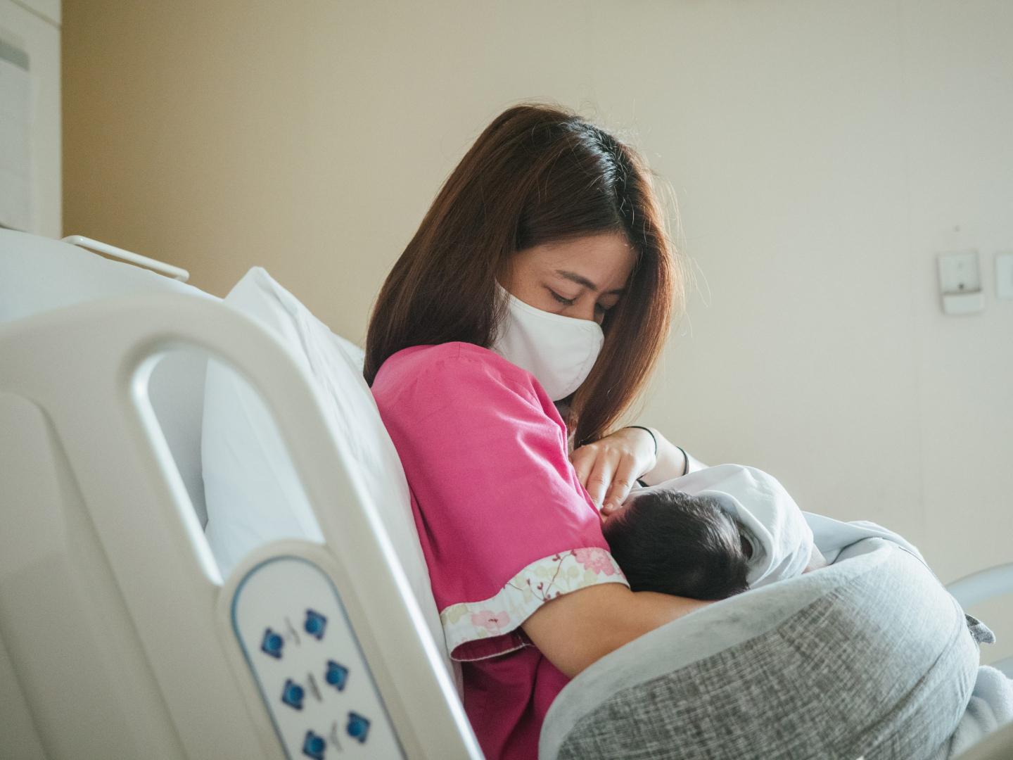 Woman in a hospital bed wearing a surgical mask and nursing her baby