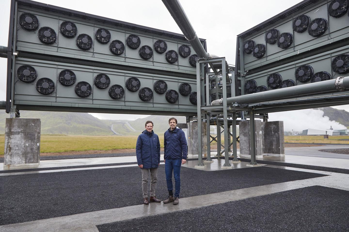 Two men standing in front of a wall of fans with the countryside in the background