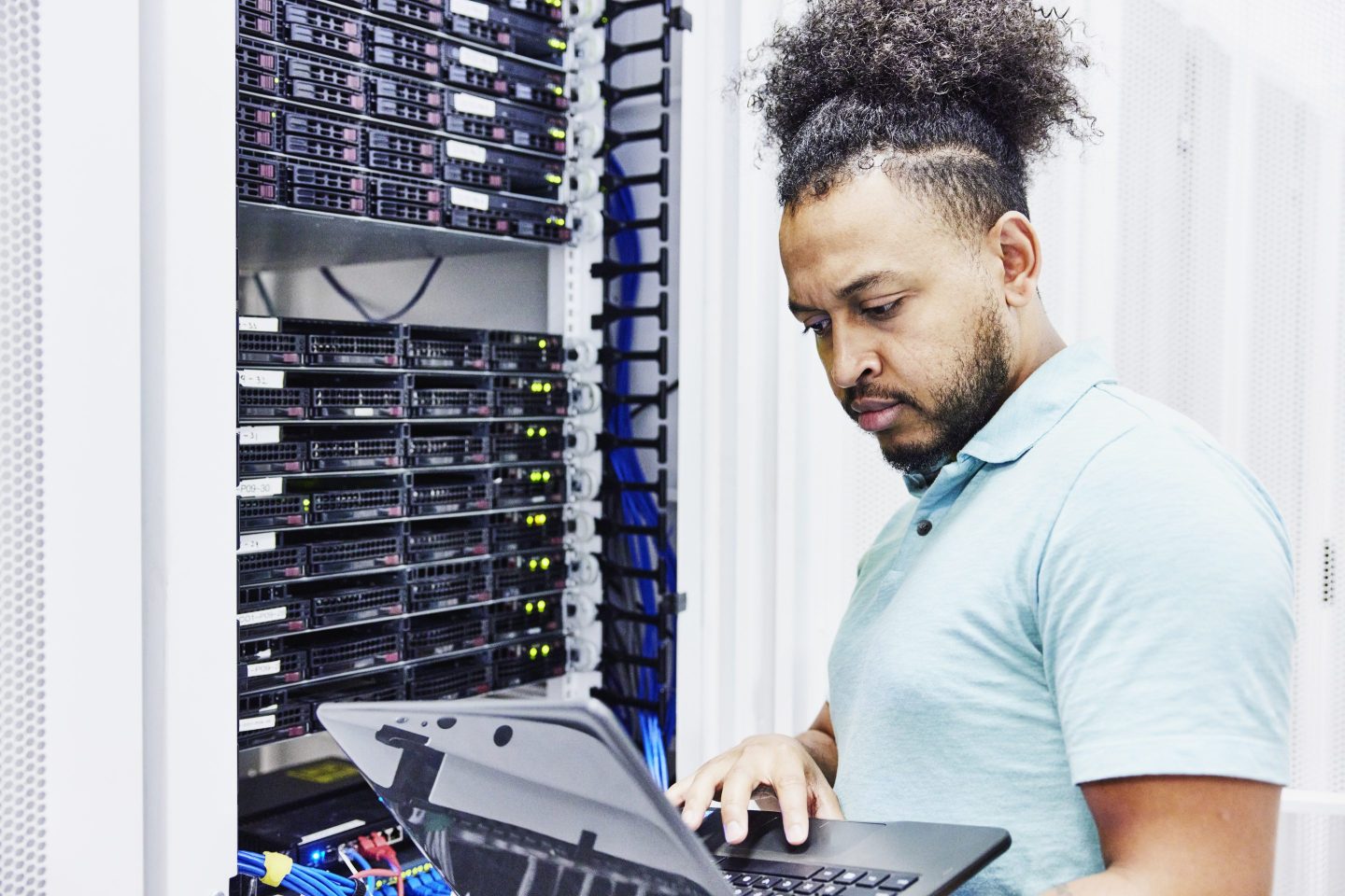 A Black man working on a laptop at a server stack.