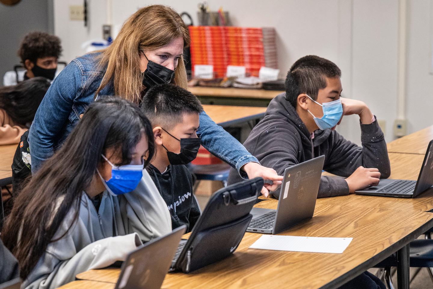 A teacher and three students wearing masks working on laptops in a classroom