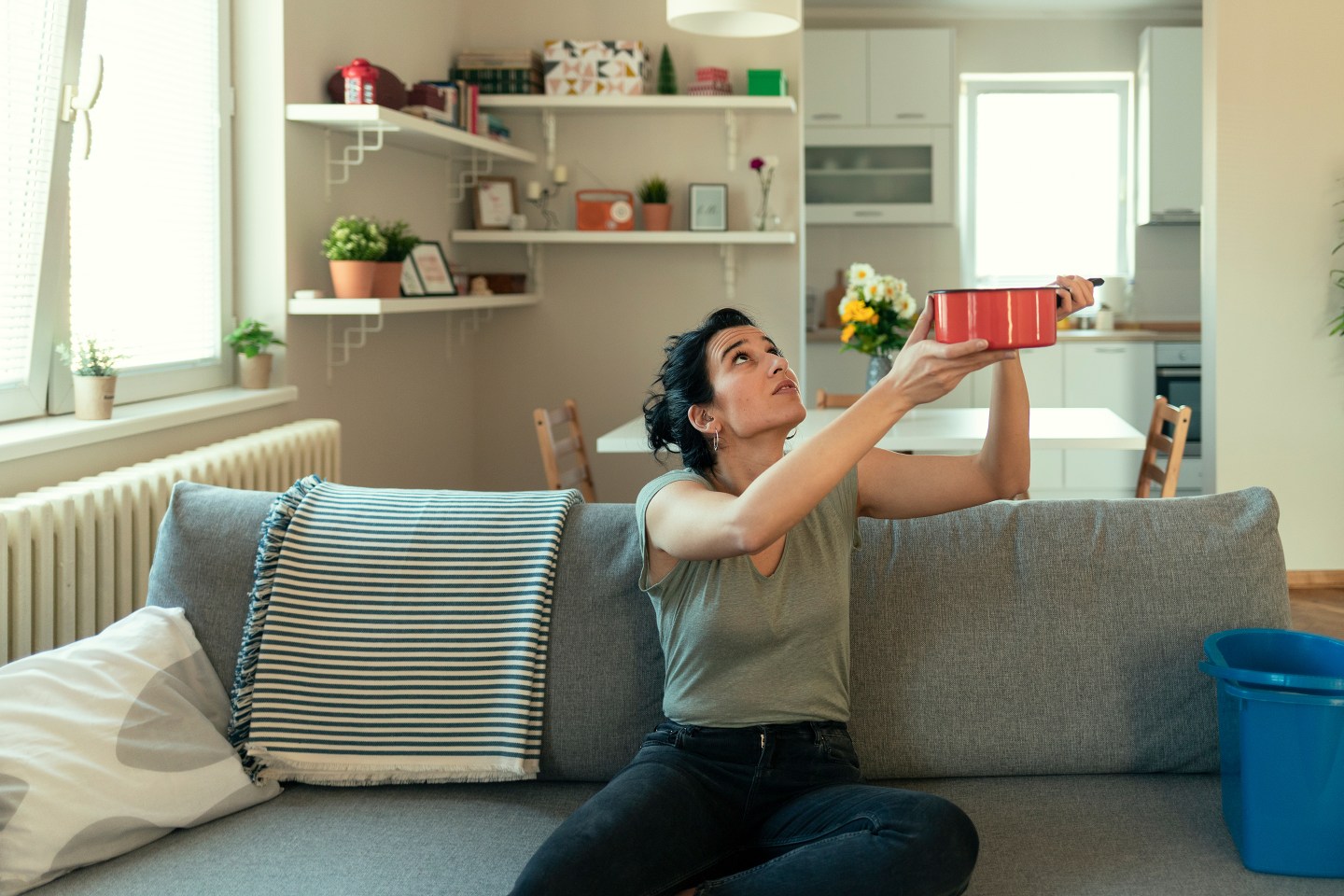 Woman holding bucket while water droplets leak from ceiling in her living room.