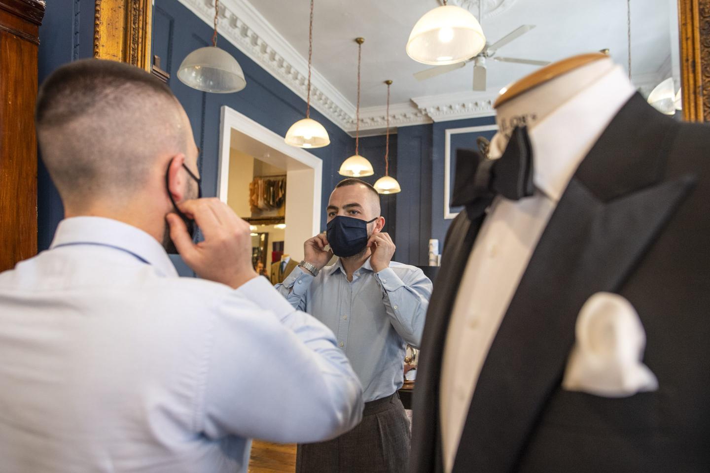 a man wearing a mask looking in the mirror. a tuxedoed mannequin in the foreground.