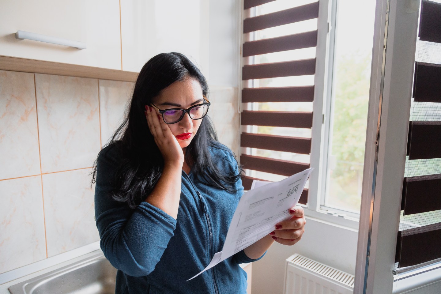 Portrait of a mid adult woman checking her energy bills at home, sitting in her bedroom. She has a worried expression and touches her face with her hand while looking at the bills. Focus on the woman while the interior architecture of the bedroom is defocused.